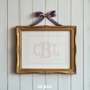 Op de afbeelding: Een ingelijste print met de initialen "CBL" in lichtroze op een witte achtergrond. De gouden lijst heeft sierlijke details en een paarse strik is bovenaan bevestigd. De print hangt aan een witte houten muur.