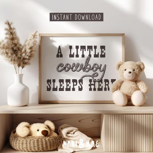 May include: A nursery print with a brown rope design that says "A little cowboy sleeps here". The print is framed and hanging on a wall above a shelf with a stuffed teddy bear and a wicker basket.