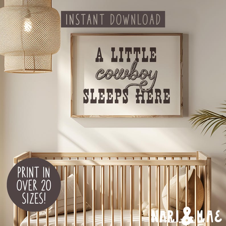 May include: A framed nursery print with the text "A little cowboy sleeps here" in black text with a rope design around the word "cowboy". The print is hanging on a white wall above a wooden crib.