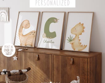 May include: Three framed prints with a dinosaur theme. The prints feature a green letter C, a yellow dinosaur, and a brown dinosaur. The prints are on a wooden shelf with the name "Charlie" written on the middle print.