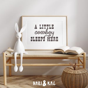 May include: A white wooden bench with a brown woven seat and a white rabbit figurine sitting on it. A framed print with the text "A little cowboy sleeps here" hangs on the wall behind the bench. A brown wicker basket sits on the floor in front of the bench.