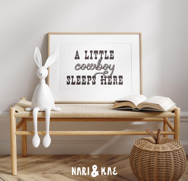 May include: A white wooden bench with a brown woven seat and a white rabbit figurine sitting on it. A framed print with the text "A little cowboy sleeps here" hangs on the wall behind the bench. A brown wicker basket sits on the floor in front of the bench.