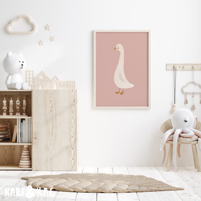 May include: A white goose illustration on a pink background in a framed print. The print is hanging on a white wall in a nursery room with a wooden bookshelf, a white teddy bear, and a white octopus plush toy.