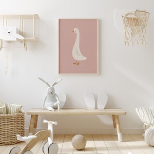 May include: A white goose illustration on a pink background, framed and hanging on a white wall in a nursery room. The room features a wooden bench, a white toy bike, a wicker basket, and a basketball hoop.