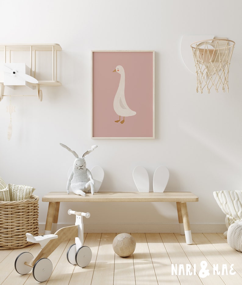 May include: A white goose illustration on a pink background, framed and hanging on a white wall in a nursery room. The room features a wooden bench, a white toy bike, a wicker basket, and a basketball hoop.