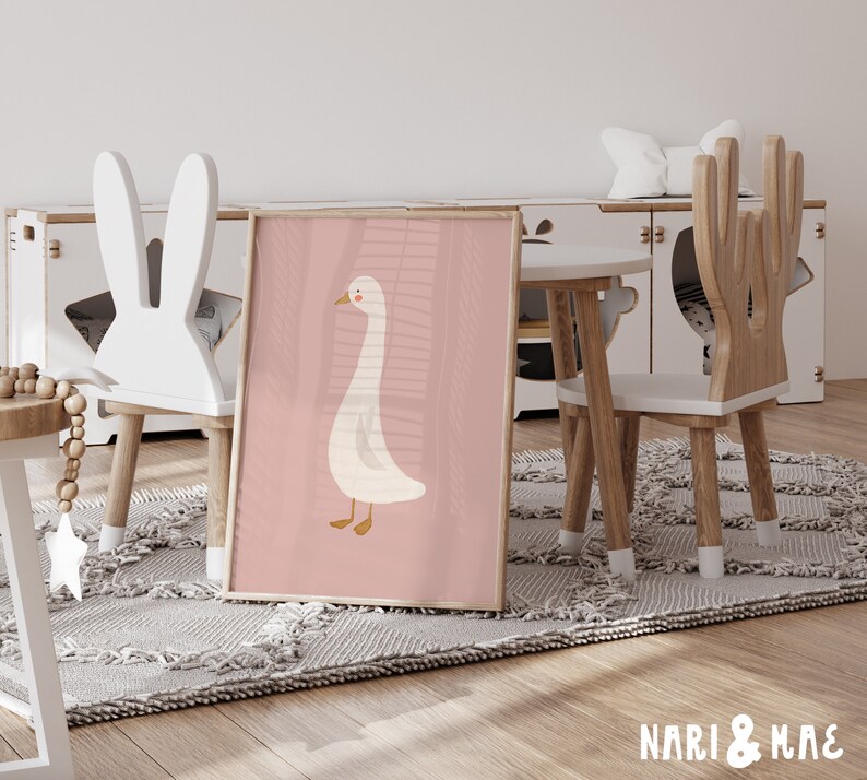 May include: A pink and white children's poster featuring a cartoon goose with a pink background. The poster is leaning against a white bunny-shaped chair in a playroom setting.