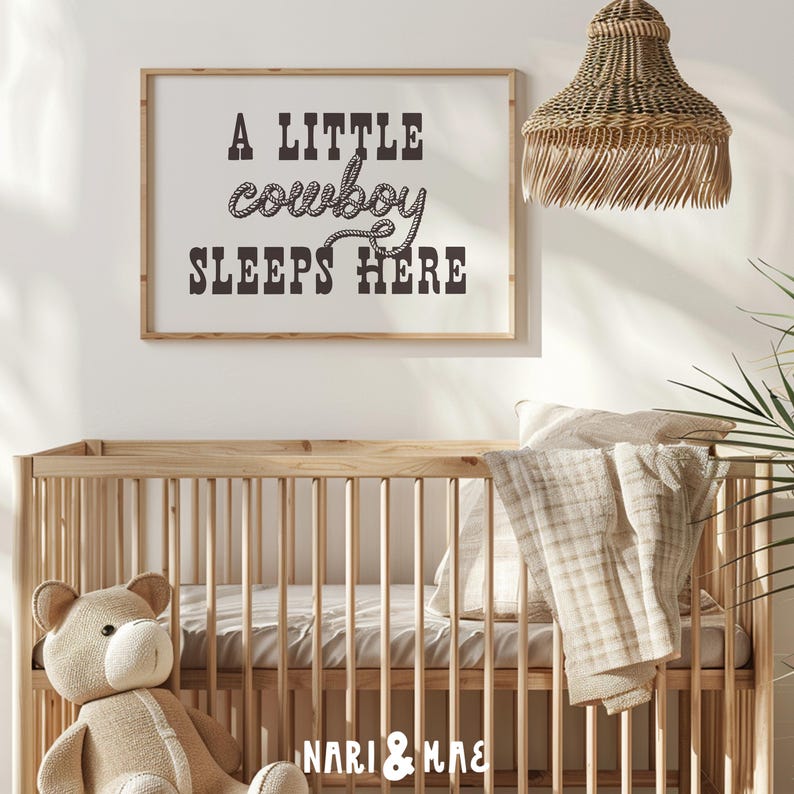 May include: A white framed print with black text that reads "A little cowboy sleeps here". The text is in a rope-like font. The print is hanging on a white wall above a wooden crib with a beige blanket and a stuffed teddy bear.