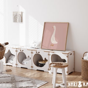 May include: White wooden storage unit with six compartments, each with a different cutout shape. The unit is decorated with a star, whale, and other shapes. A white stool with a wooden top sits in front of the unit. A framed print of a white goose on a pink background hangs on the wall above the unit. The text "NARI & MAE" is visible in the bottom right corner of the image.