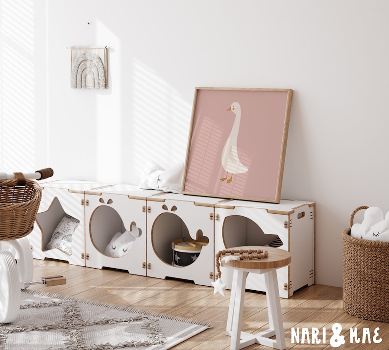 May include: White wooden storage unit with six compartments, each with a different cutout shape. The unit is decorated with a star, whale, and other shapes. A white stool with a wooden top sits in front of the unit. A framed print of a white goose on a pink background hangs on the wall above the unit. The text "NARI & MAE" is visible in the bottom right corner of the image.