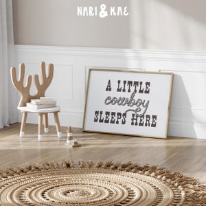 May include: A gold framed print with the text "A little cowboy sleeps here" in black lettering with a rope design around the word "cowboy". The print is hanging on a light beige wall above a light brown woven rug with fringe.