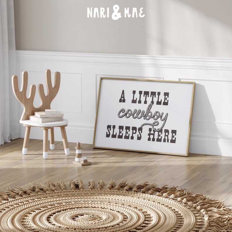 May include: A gold framed print with the text "A little cowboy sleeps here" in black lettering with a rope design around the word "cowboy". The print is hanging on a light beige wall above a light brown woven rug with fringe.