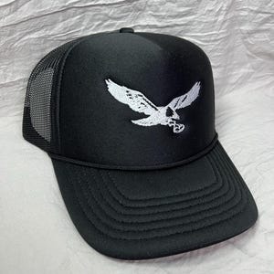 May include: Black trucker hat with a white embroidered eagle design. The hat has a mesh back and a curved brim.