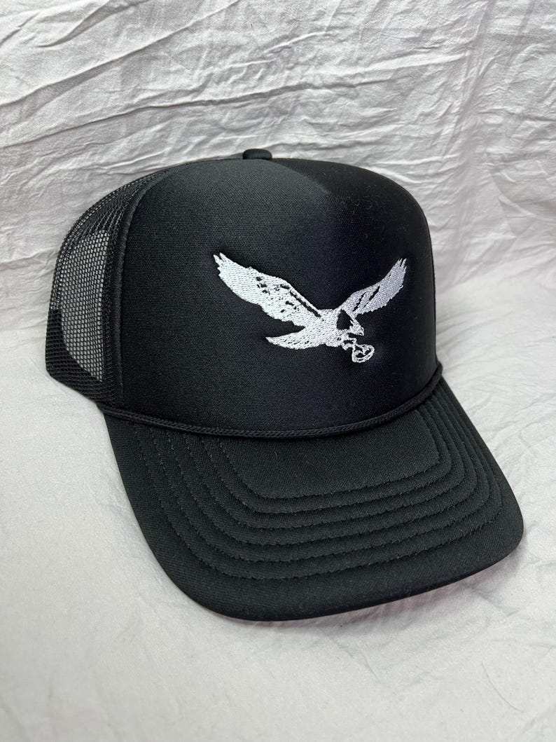 May include: Black trucker hat with a white embroidered eagle design. The hat has a mesh back and a curved brim.