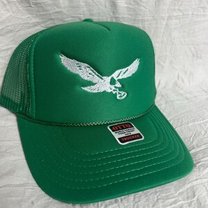 May include: Green trucker hat with a white embroidered eagle design. The hat has a mesh back and a label that reads "OTTO WORLD FAMOUS FOAM FRONT OWN TRUCKER".