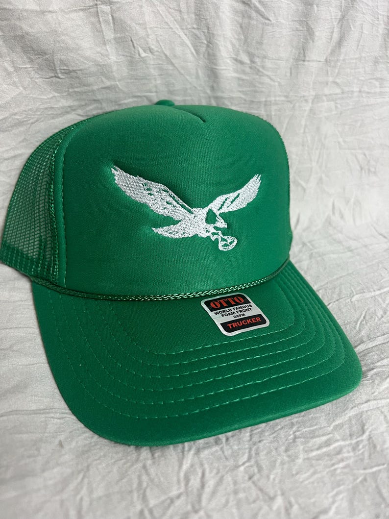 May include: Green trucker hat with a white embroidered eagle design. The hat has a mesh back and a label that reads "OTTO WORLD FAMOUS FOAM FRONT OWN TRUCKER".