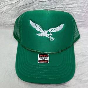 May include: Green trucker hat with a white embroidered eagle design. The hat has a mesh back and a label that reads "OTTO WORLD FAMOUS FOAM FRONT OSFM TRUCKER".