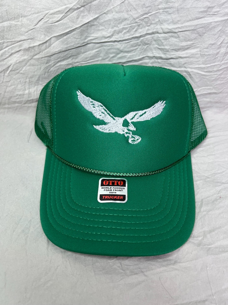 May include: Green trucker hat with a white embroidered eagle design. The hat has a mesh back and a label that reads "OTTO WORLD FAMOUS FOAM FRONT OSFM TRUCKER".
