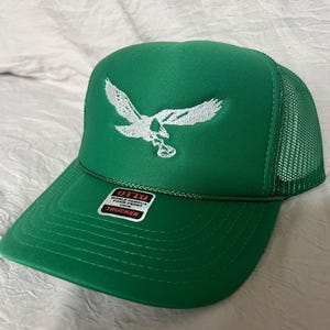 May include: Green trucker hat with a white embroidered eagle design. The hat has a mesh back and a label that reads "OTTO WORLD FAMOUS FOAM FRONT COVER TRUCKER".