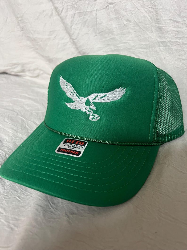 May include: Green trucker hat with a white embroidered eagle design. The hat has a mesh back and a label that reads "OTTO WORLD FAMOUS FOAM FRONT COVER TRUCKER".