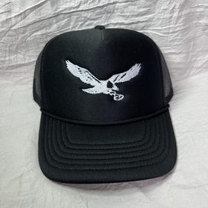 May include: A black trucker hat with a white embroidered eagle design. The hat has a mesh back and a black brim.