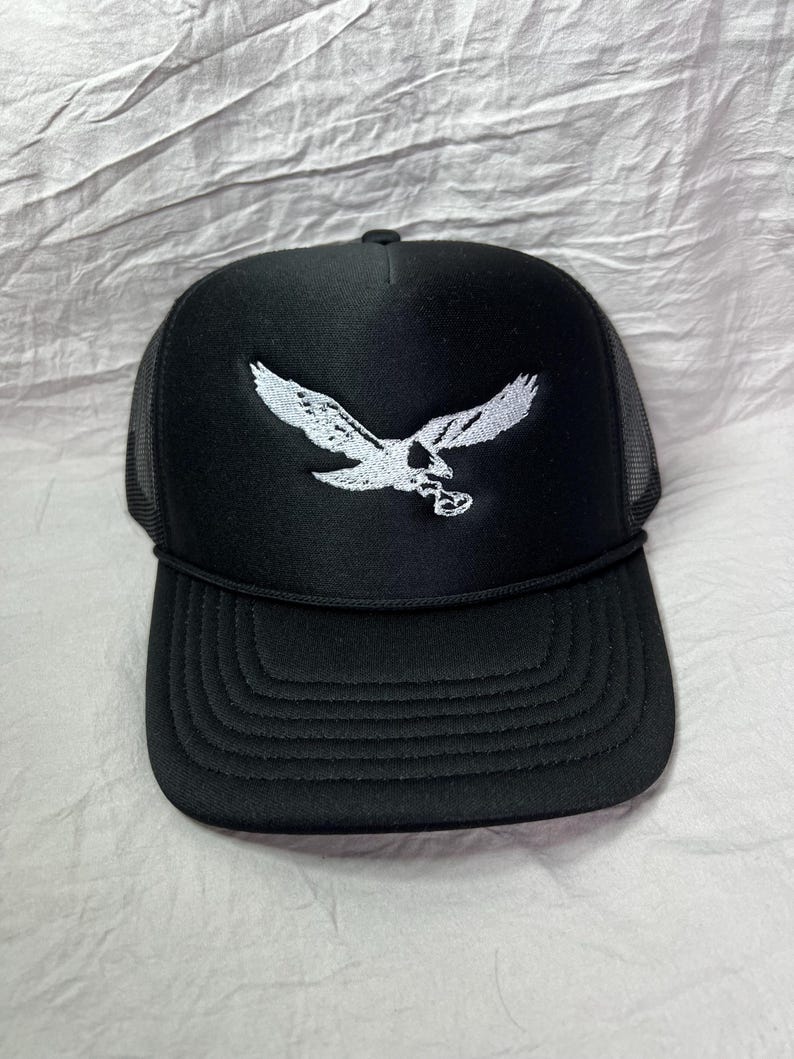 May include: A black trucker hat with a white embroidered eagle design. The hat has a mesh back and a black brim.