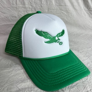 May include: A white and green trucker hat with a green embroidered eagle on the front. The hat has a mesh back and a green brim.
