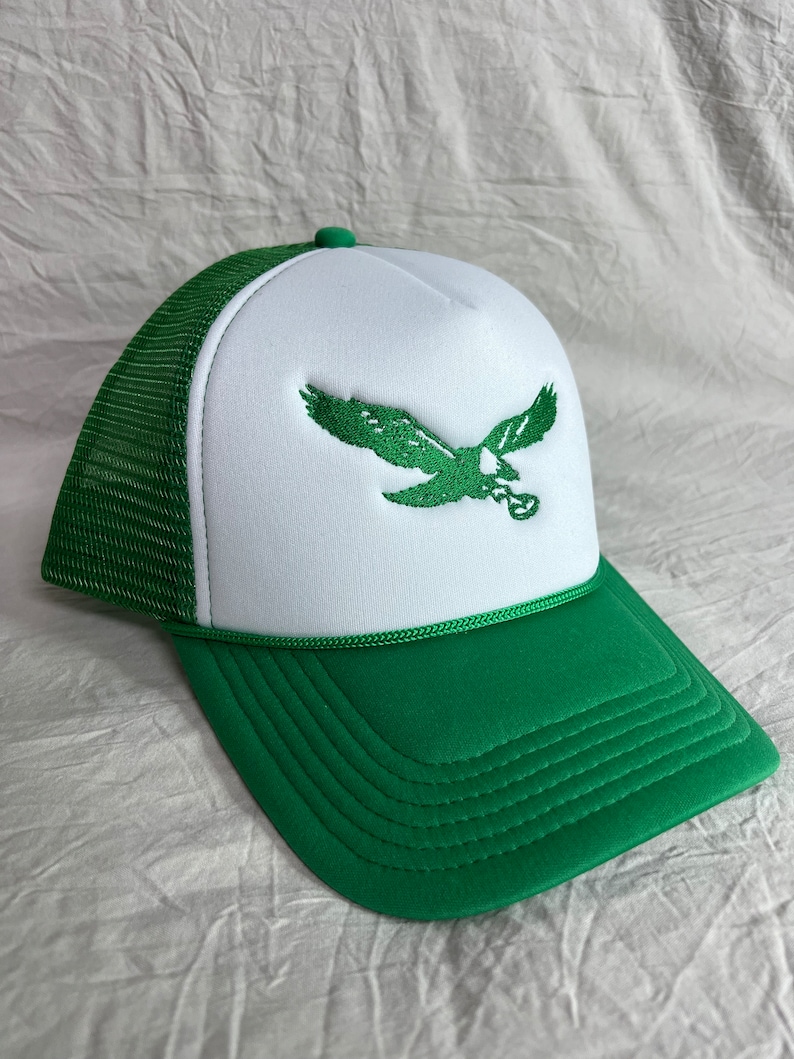May include: A white and green trucker hat with a green embroidered eagle on the front. The hat has a mesh back and a green brim.