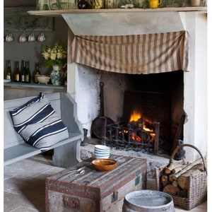 May include: A rustic fireplace with a striped fabric covering the mantel, a wood-burning fire, and a wooden bench with a striped pillow. A wooden trunk coffee table sits in front of the fireplace with a bowl and two mugs on top. A wooden barrel sits next to the trunk.