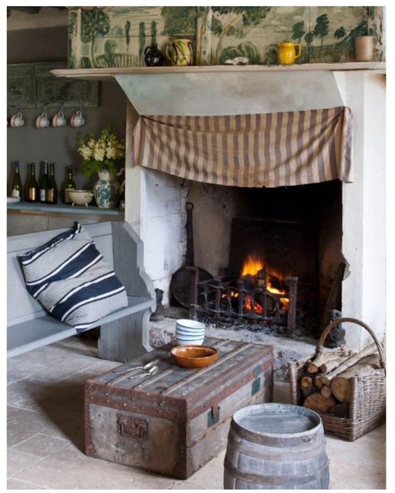 May include: A rustic fireplace with a striped fabric covering the mantel, a wood-burning fire, and a wooden bench with a striped pillow. A wooden trunk coffee table sits in front of the fireplace with a bowl and two mugs on top. A wooden barrel sits next to the trunk.