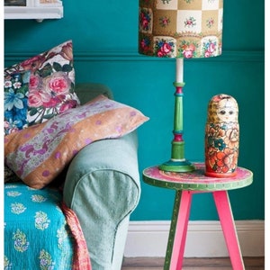May include: A teal blue wall with a green lamp with a floral patterned lampshade. The lamp is on a small pink and green table with a painted design. A Russian nesting doll is on the table.