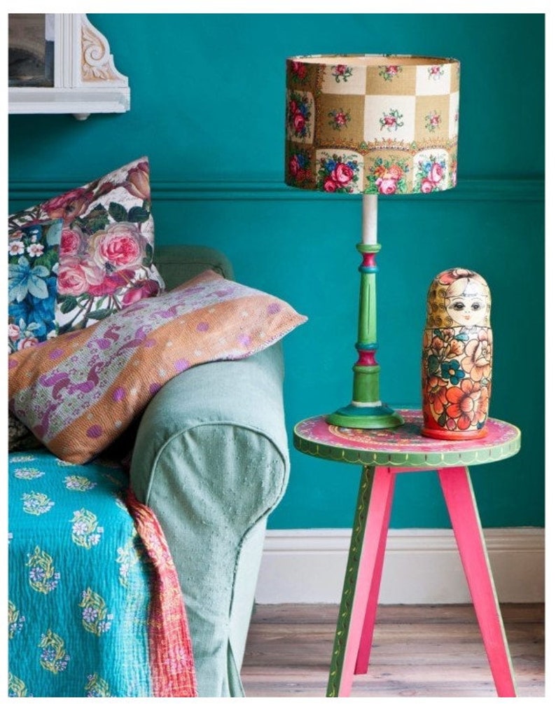 May include: A teal blue wall with a green lamp with a floral patterned lampshade. The lamp is on a small pink and green table with a painted design. A Russian nesting doll is on the table.