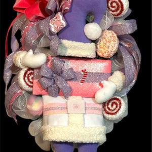 May include: A purple and white Christmas wreath featuring a Santa figure holding a pink gift box with a purple bow. The wreath is decorated with white, pink, and red candy canes and other festive embellishments.