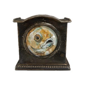 May include: A small, antique-style clock with a fish illustration. The clock has a square, dark metal frame with decorative details. The fish is painted in watercolor with shades of orange, brown, and blue.