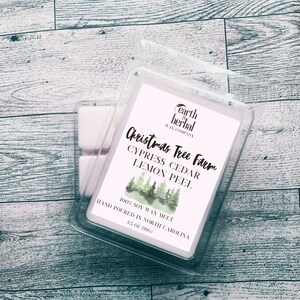 Könnte beinhalten: Ein weißer Wachsschmelzer mit einer Aquarell-Illustration von Kiefern. Das Etikett lautet "earth & herbal wax company" und "Christmas Tree Farm Cypress Cedar Lemon Peel 100% Soy Wax Melt Hand Poured in North Carolina 3.5 oz (99g)"