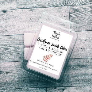 Puede incluir: Un derretimiento de cera blanco con una etiqueta de "Christmas Snack Cakes". La etiqueta presenta una ilustración festiva de un pastel de queso crema de almendras y vainilla. El texto en la etiqueta dice "earth & herbal wax company", "Christmas Snack Cakes", "Vanilla Almond Cream Cheese", "100% Soy Wax Melt", "Hand Poured in North Carolina", y "3.5 oz (99g)".