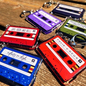 Puede incluir: Una colección de llaveros de cintas de cassette de colores. Cada llavero presenta un color y diseño diferente, con textos como "Save Rock and Roll" e "Infinity on High". Los llaveros tienen anillos plateados.