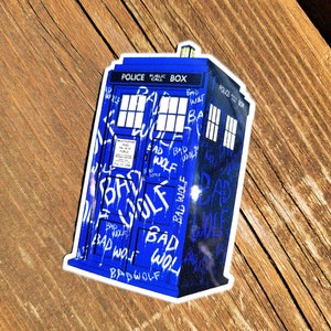 May include: A blue sticker depicting a police box, with the words "POLICE PUBLIC CALL BOX" at the top. The box is covered in white graffiti that reads "BAD WOLF". The sticker has a white border.