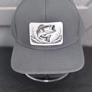 May include: A gray baseball cap with a curved brim. The cap features a rectangular patch with a black and white illustration of a fish. The fish is open-mouthed, surrounded by stylized branches.