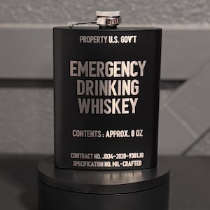 May include: Black flask with the text "EMERGENCY DRINKING WHISKEY" and "PROPERTY U.S. GOV'T". The flask holds approximately 8 oz. of liquid and has a silver cap. The flask is on a black stand.