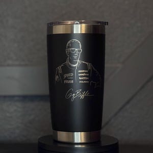 May include: Black stainless steel tumbler with a silver rim and lid. The tumbler features an etched portrait of Greg Biffle, along with his signature and sponsor logos. The tumbler is designed to keep beverages hot or cold.