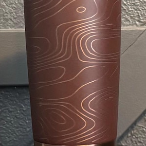 May include: A stainless steel tumbler with a copper base, rim, and lid. The body is dark brown with a topographic map design in a lighter copper colour. The tumbler sits on a black base.