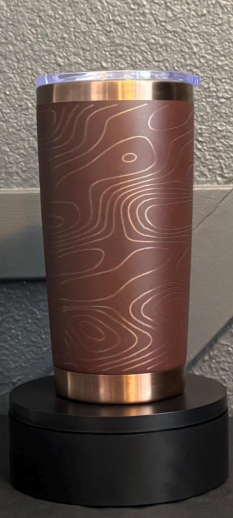 May include: A stainless steel tumbler with a copper base, rim, and lid. The body is dark brown with a topographic map design in a lighter copper colour. The tumbler sits on a black base.