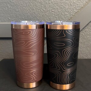 May include: Two insulated tumblers with copper-coloured accents. One is brown with a topographic map design, the other black with a similar map pattern. Both have clear lids.