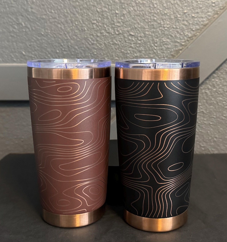 May include: Two insulated tumblers with copper-coloured accents. One is brown with a topographic map design, the other black with a similar map pattern. Both have clear lids.