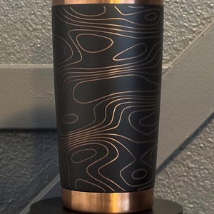 May include: A black tumbler with a copper-coloured rim and base. The tumbler features a topographic map design in a copper colour. The tumbler is cylindrical and stands on a black base.