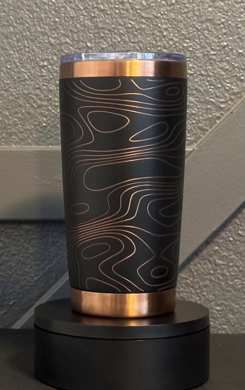 May include: A black tumbler with a copper-coloured rim and base. The tumbler features a topographic map design in a copper colour. The tumbler is cylindrical and stands on a black base.
