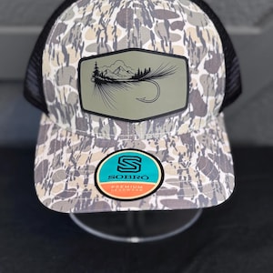 May include: A camouflage-patterned trucker hat with a black mesh back and a curved brim. The front features a patch with a mountain and fly fishing hook design. The hat also has a circular sticker with the brand name "SOBRO" and the words "PREMIUM HEADWEAR".