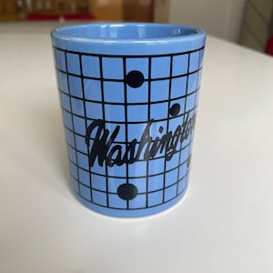 May include: A blue ceramic mug with a black grid pattern and the word "Washington" written in a black cursive font. There are black circles on the grid.