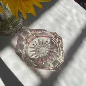 May include: A clear glass ashtray with a starburst design and a pink rim. The ashtray is octagonal in shape and has a slightly raised edge.