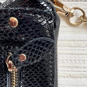 Peut inclure: Gros plan sur un portefeuille noir textur&eacute; avec un motif en forme de reptile. Le portefeuille est orn&eacute; d'&eacute;l&eacute;ments dor&eacute;s, notamment une fermeture &eacute;clair, des rivets et un fermoir &agrave; cha&icirc;ne. Le design est d&eacute;taill&eacute; et le mat&eacute;riau semble &ecirc;tre du cuir.